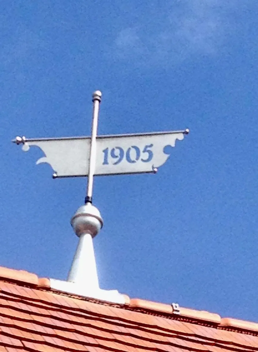 Ein weißer Wetterfahne auf einem orangefarbenen Ziegeldach mit der Jahreszahl "1905" vor blauem Himmel.