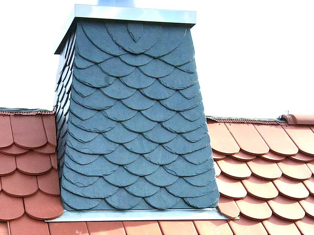 Schornstein mit Schieferverkleidung auf einem Dach mit roten Ziegeln.