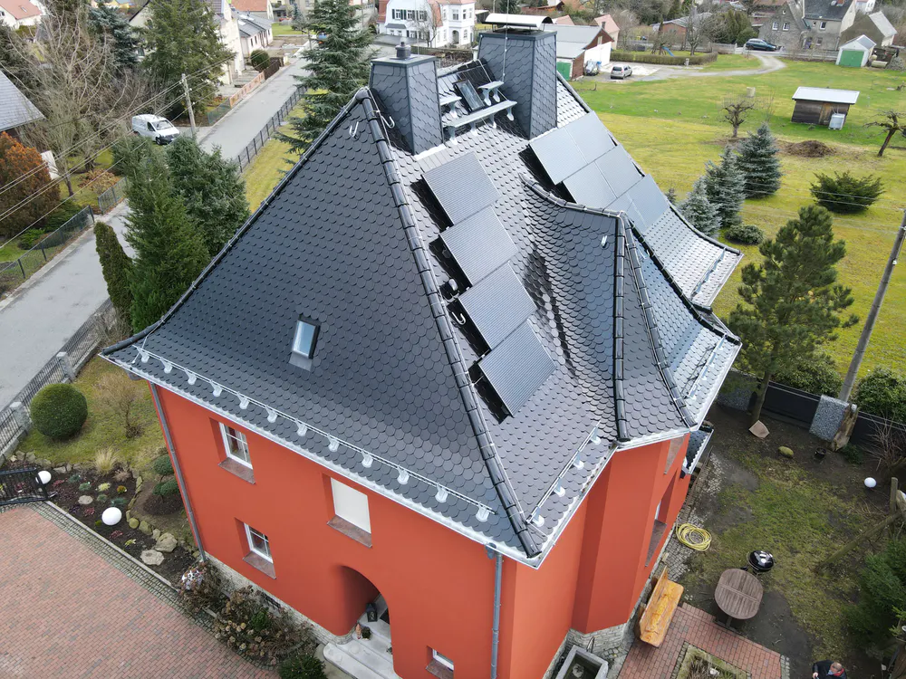 Rotes Haus mit schwarzem Dach und Photovoltaikmodulen in einer Wohnsiedlung.