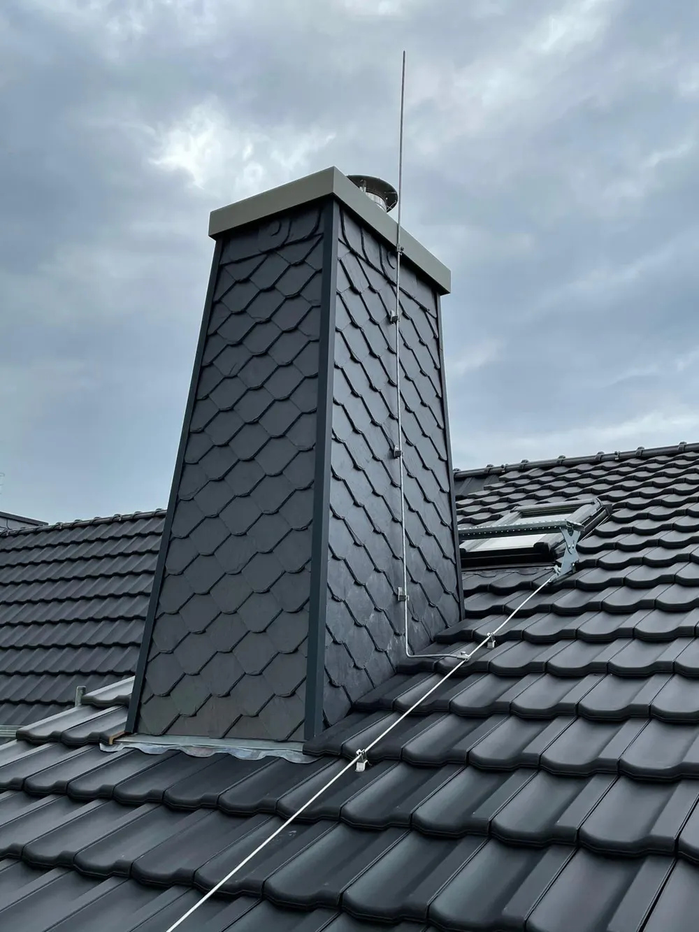 Dunkelgrauer Schornstein mit Schieferverkleidung und Blitzableiter auf einem schwarzen Ziegeldach unter bewölktem Himmel.