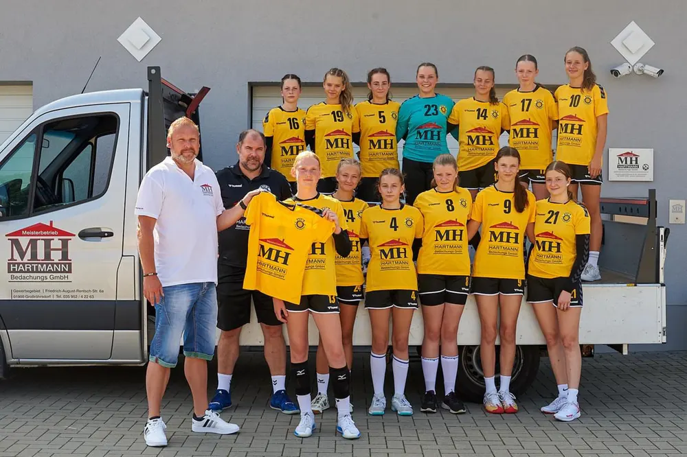 Handballteam in gelben Trikots mit "MH HARTMANN" Logo, stehend vor einem Transporter und einem Gebäude.
