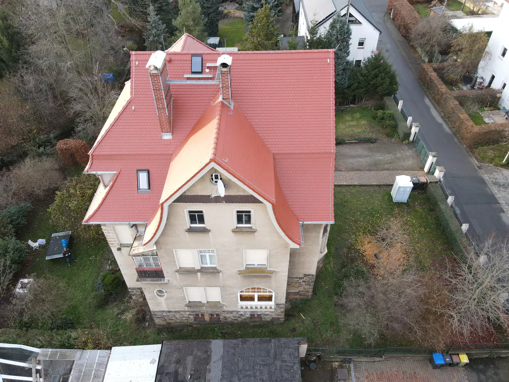 Historisches Haus mit rotem Ziegeldach, beigefarbenen Wänden und mehreren Fenstern, umgeben von Bäumen und einer Straße.