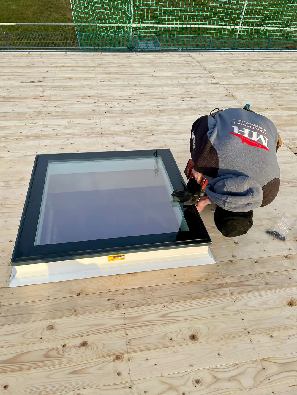 Ein Arbeiter befestigt ein Dachfenster auf einer Holzterrasse mit einem grünen Netz im Hintergrund.