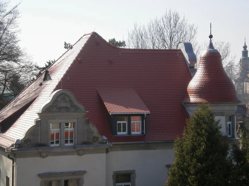 Rotes Ziegeldach eines Gebäudes mit Gauben und einem runden Turm, umgeben von Bäumen.