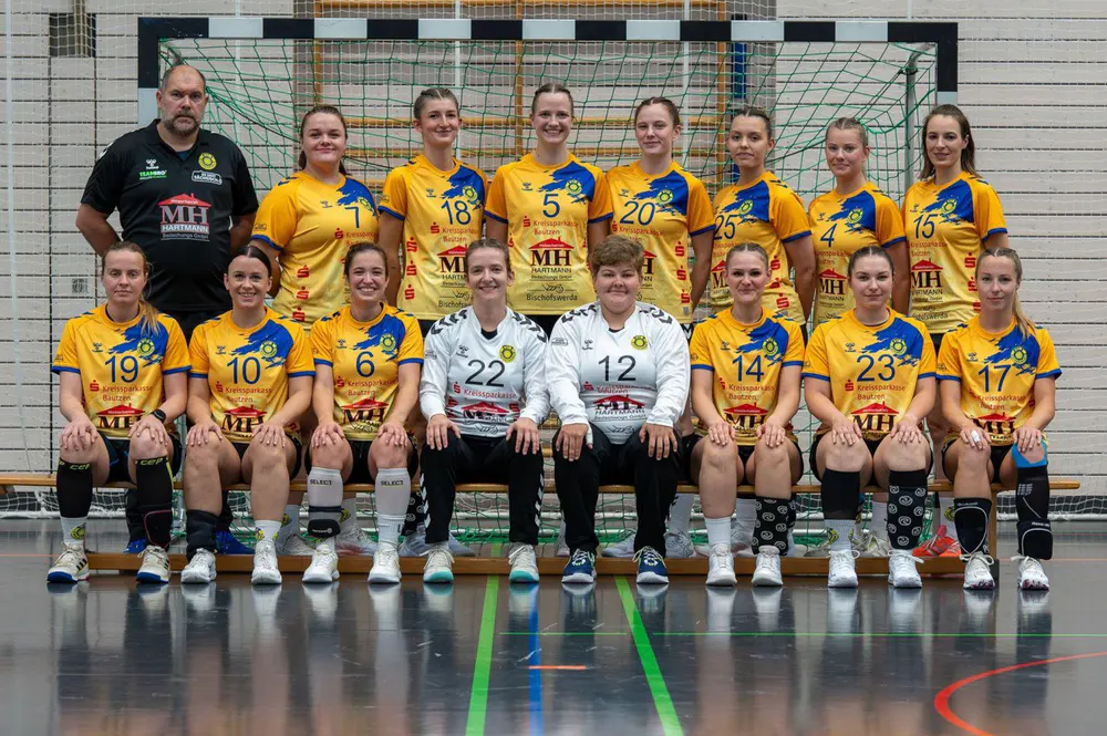 Ein Handballteam von 16 Frauen und einem Mann posiert in gelb-blauen Trikots in einer Sporthalle.