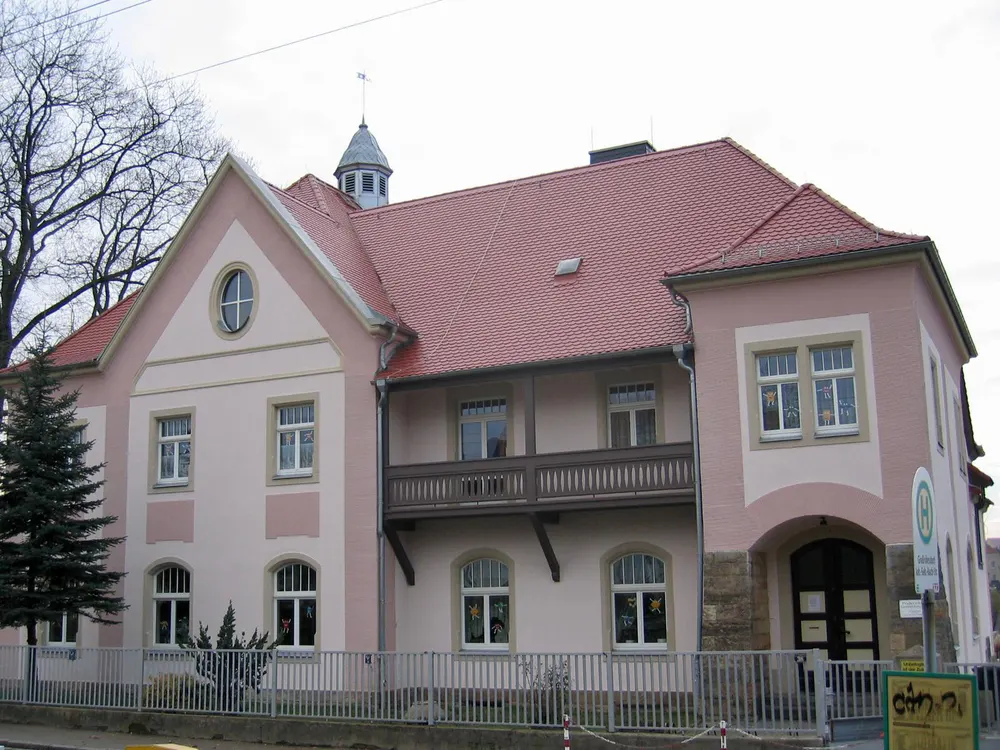 Rosa Gebäude mit rotem Ziegeldach, Dachreiter, Holzbalkon und hohen Fenstern unter bewölktem Himmel.