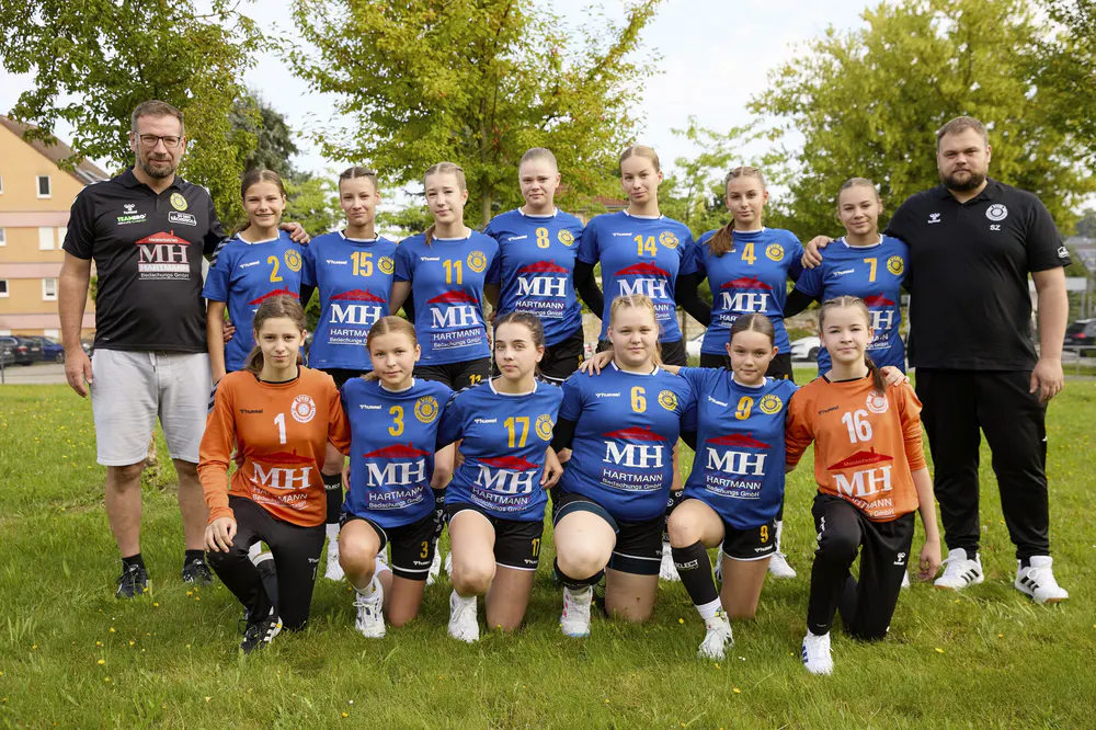 Handballteam von 14 Mädchen in blauen und orangefarbenen Trikots mit zwei Trainern auf einer Wiese.