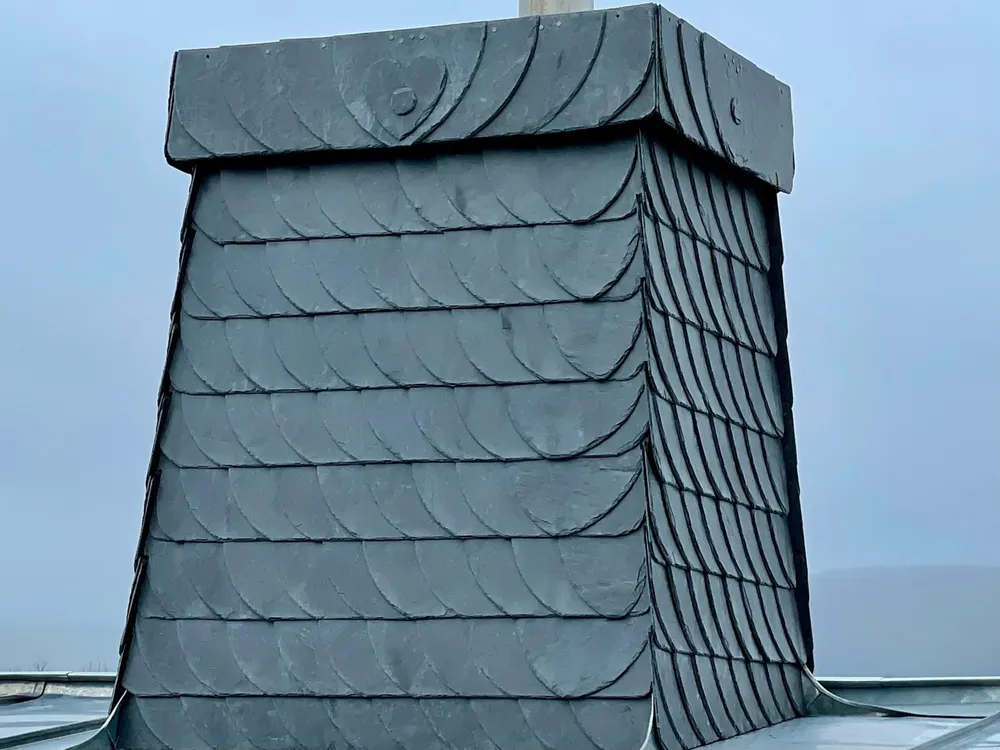 Schornstein mit dunklen Schieferplatten bedeckt, die in Fischschuppenmuster angeordnet sind, vor grauem Himmel.