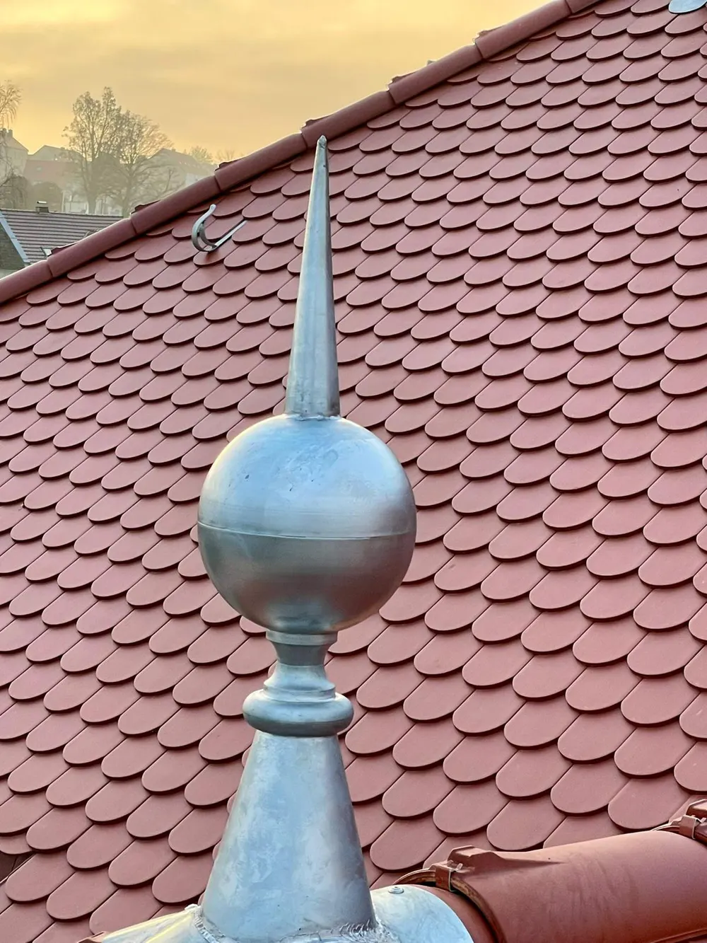 Spitze mit Kugel und kegelförmigem Sockel auf einem Dach mit roten Biberschwanzziegeln.