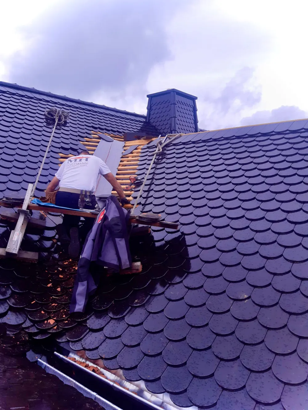 Dachdecker repariert ein schwarzes Schuppendach mit Schornstein unter bewölktem Himmel.