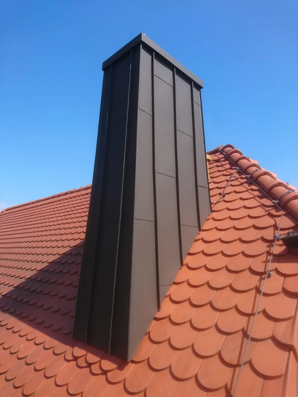 Brauner Schornstein auf einem roten Ziegeldach unter blauem Himmel.