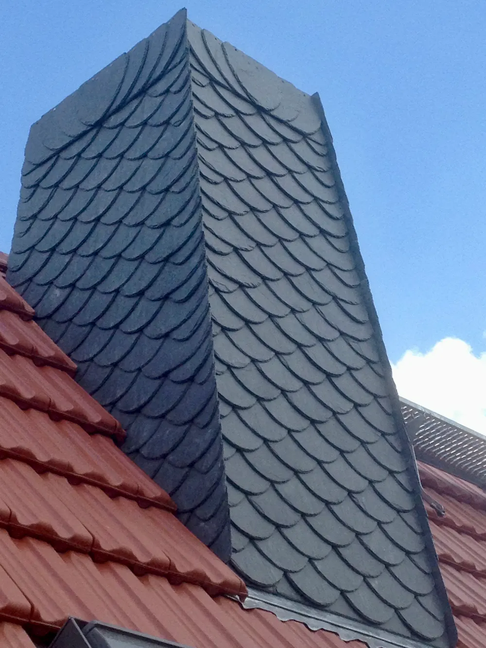 Schornstein mit dunklen Schindeln aus Schiefer und rotem Ziegeldach unter blauem Himmel.