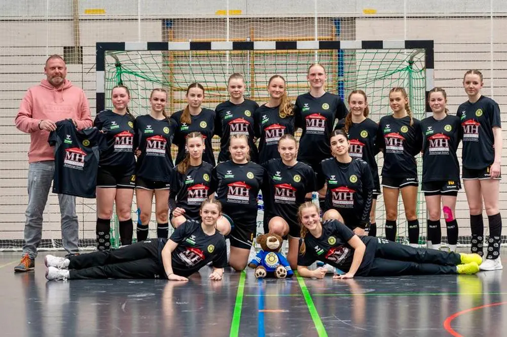 Eine Handballmannschaft von 18 Frauen und einem Mann posiert in schwarzen Trikots mit "MH" Logo vor einem Tor.