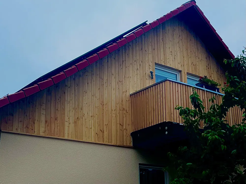 Hausfassade mit Holzverkleidung, rotem Ziegeldach und kleinem Holzbalkon.