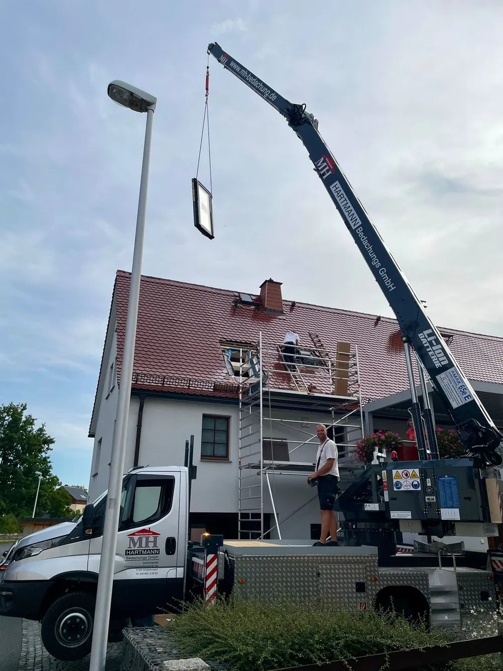 Ein großer Kran hebt ein Dachfenster über ein Haus mit rotem Dach, während Arbeiter auf dem Gerüst stehen.
