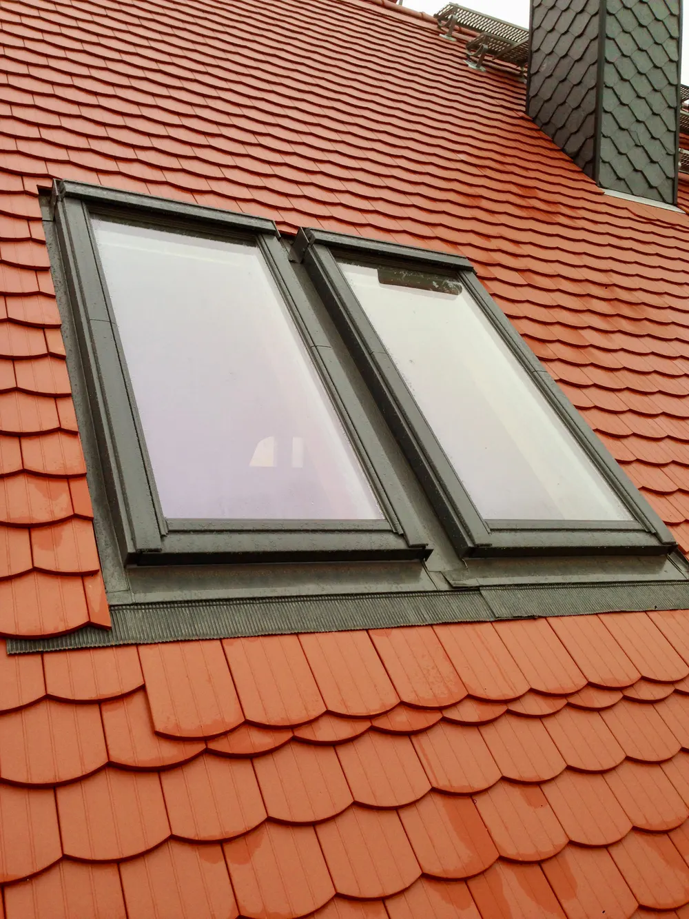 Zwei Dachfenster in einem roten Ziegeldach, ein Schornstein ist im Hintergrund sichtbar.