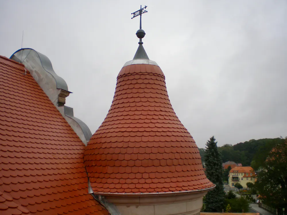 Rotes Ziegeldach mit rundem Turm und Wetterfahne auf einem Gebäude vor bewölktem Himmel.