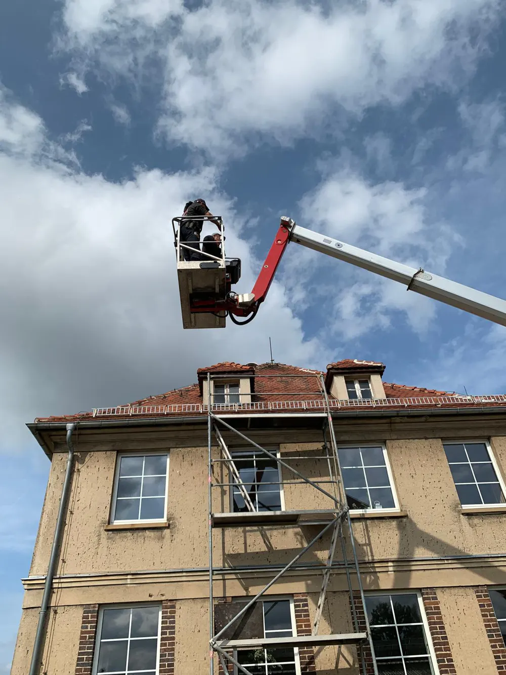 Arbeiter in einem Hubsteiger über einem Gebäude mit Gerüst und roten Ziegeldach.