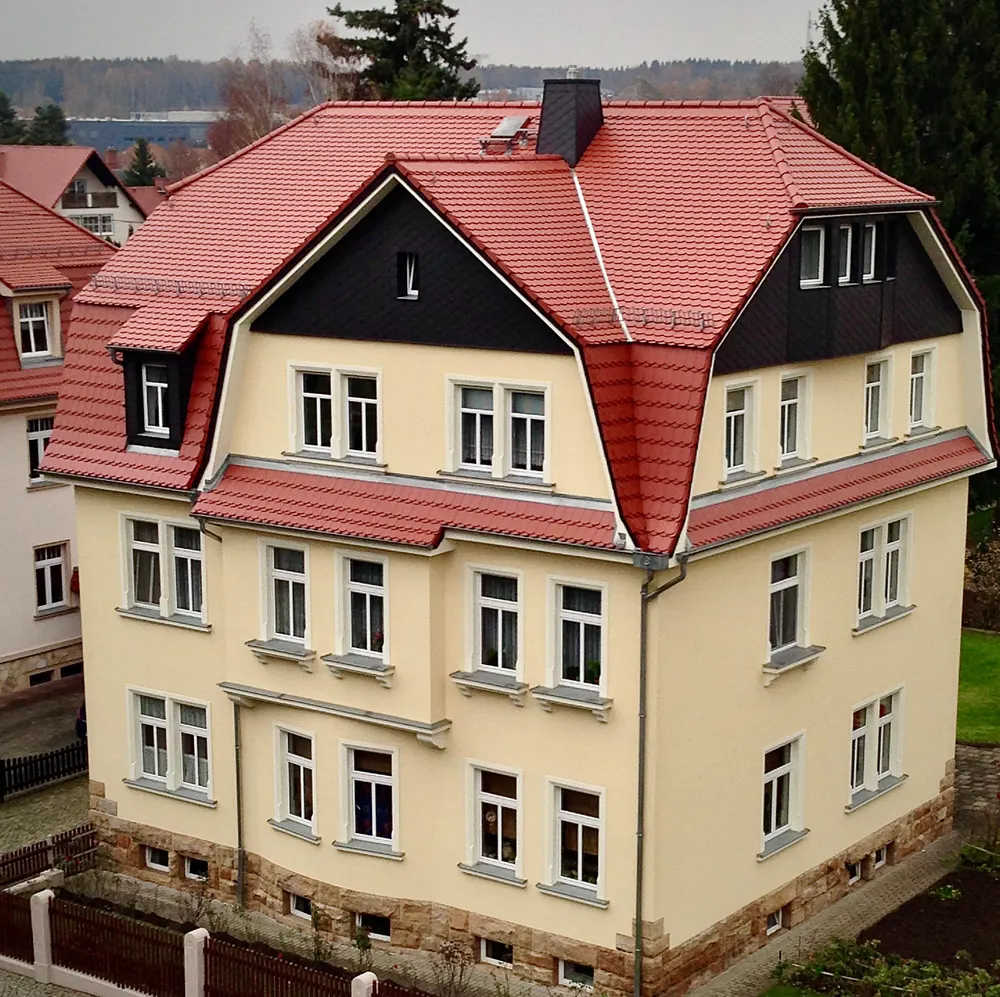 Mehrstöckiges, hellgelbes Haus mit rotem Ziegeldach, schwarzen Giebeln und vielen weißen Fenstern.