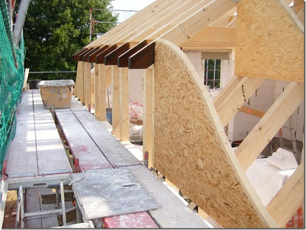 Dachstuhl im Bau mit freiliegenden Holzsparren und Schalung, ein Fenster ist sichtbar.