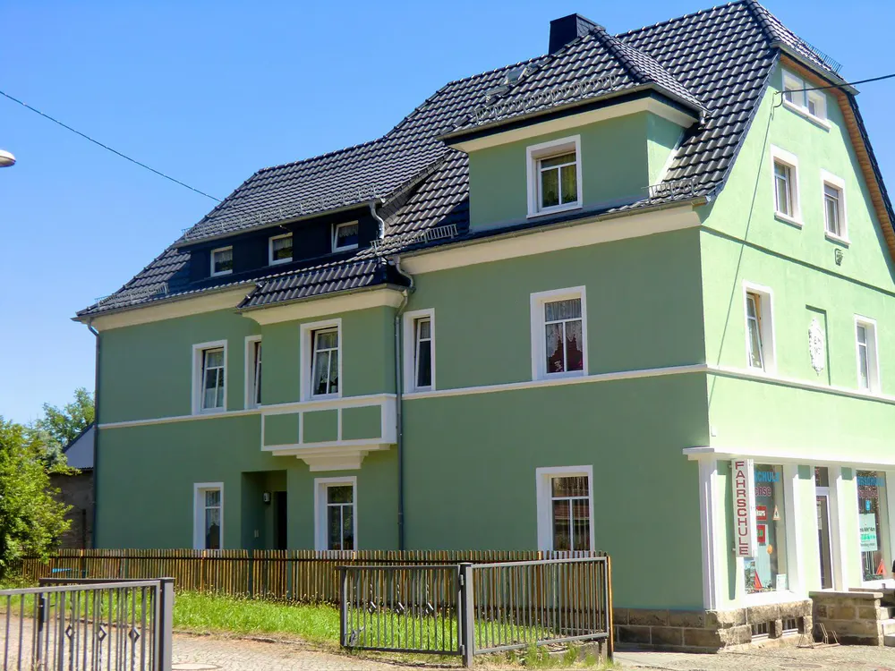 Grünes Haus mit dunklem Dach und weißen Fensterrahmen, im Vordergrund ein Metallzaun.