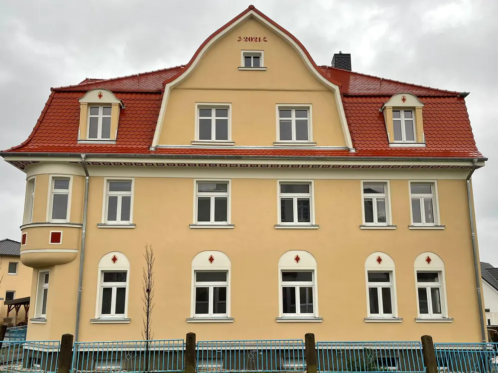 Gelbes Haus mit rotem Ziegeldach, weißen Fenstern und einem Erker, davor ein blauer Zaun.