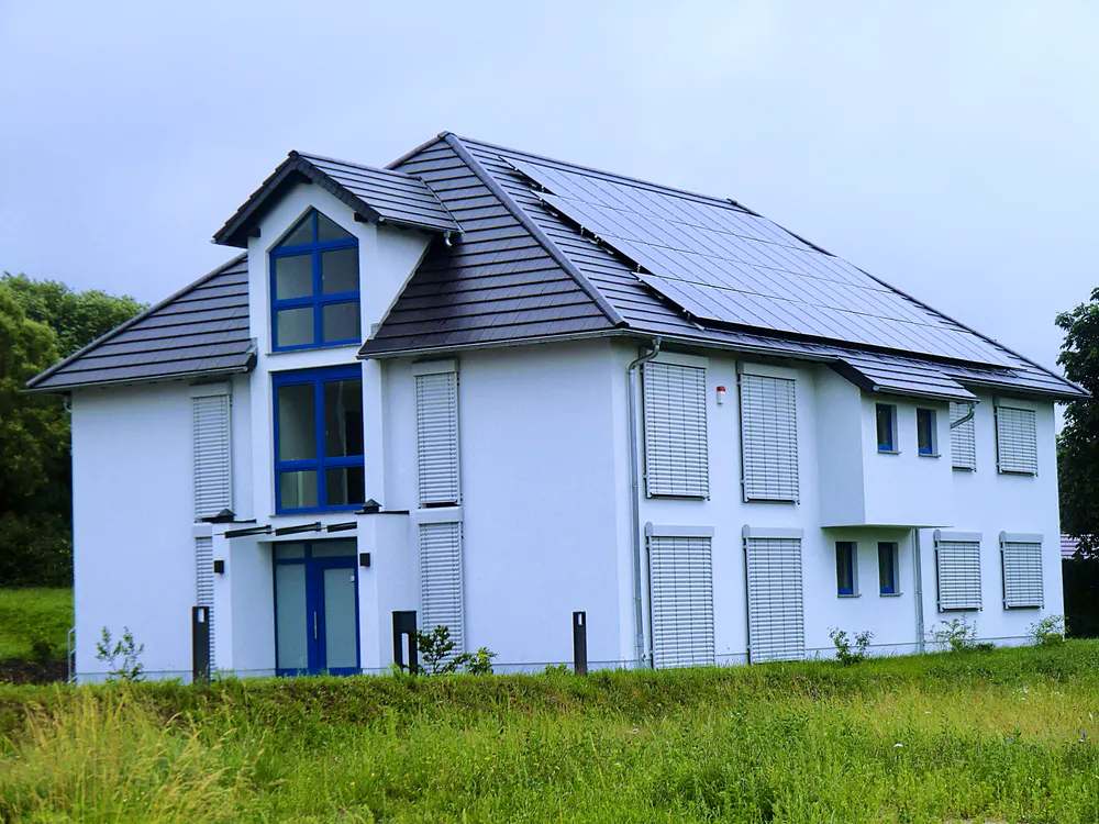 Weißes Einfamilienhaus mit Solaranlage auf dem Dach und geschlossenen Rollläden, umgeben von grünen Feldern.