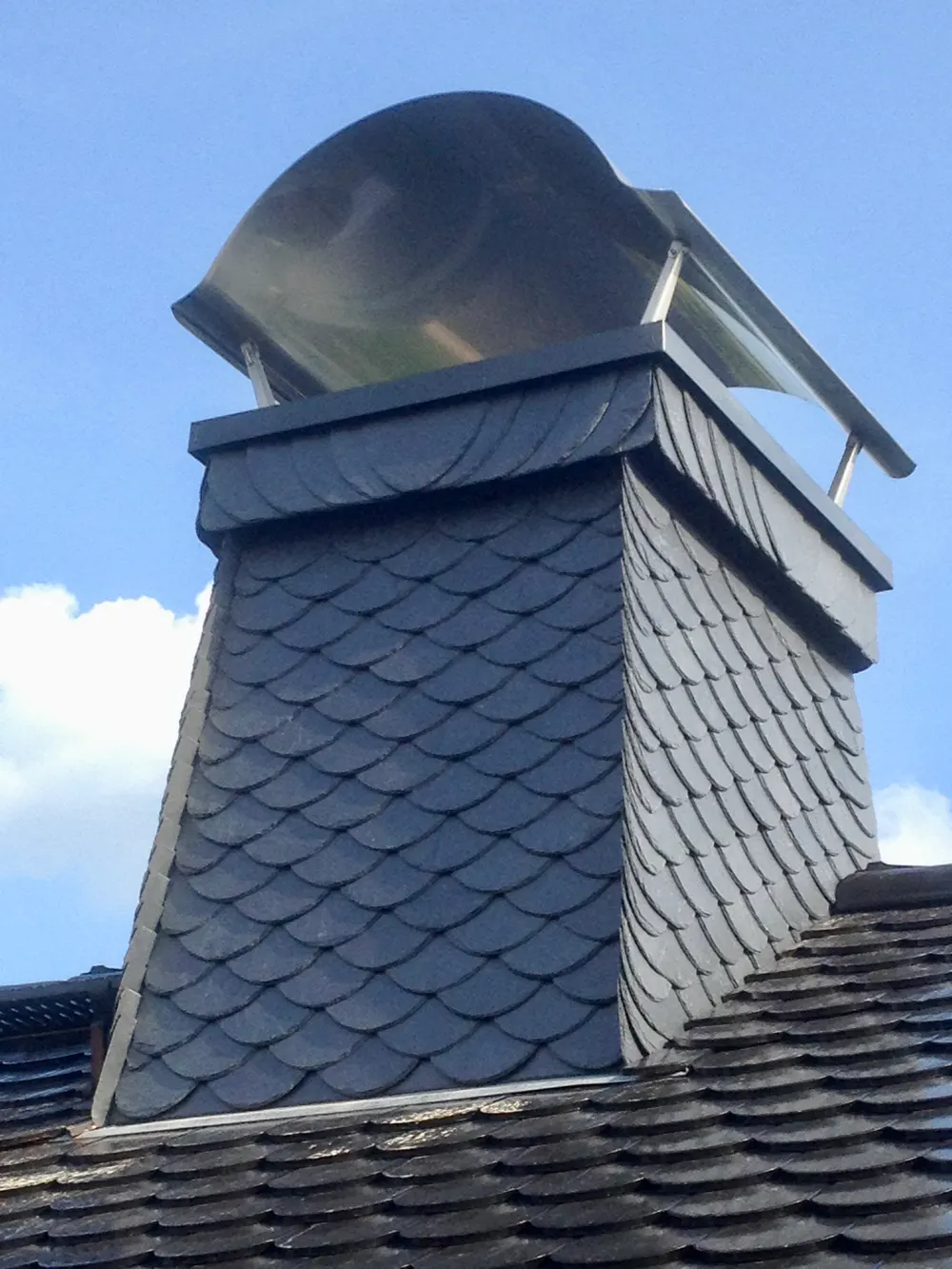 Schornstein mit Schieferverkleidung und Metallhaube auf einem Ziegeldach unter blauem Himmel.
