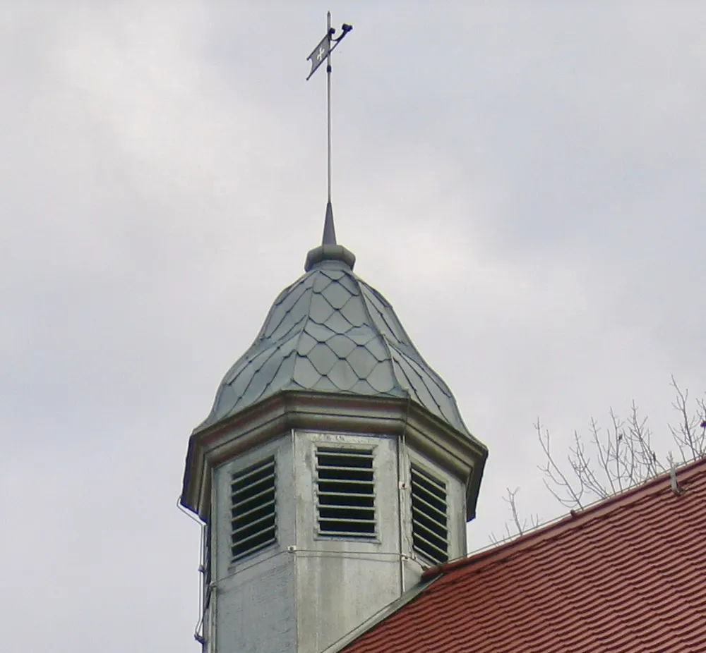Spitzer, schiefergedeckter Turm mit Lamellenfenstern und Wetterfahne auf einem roten Ziegeldach.