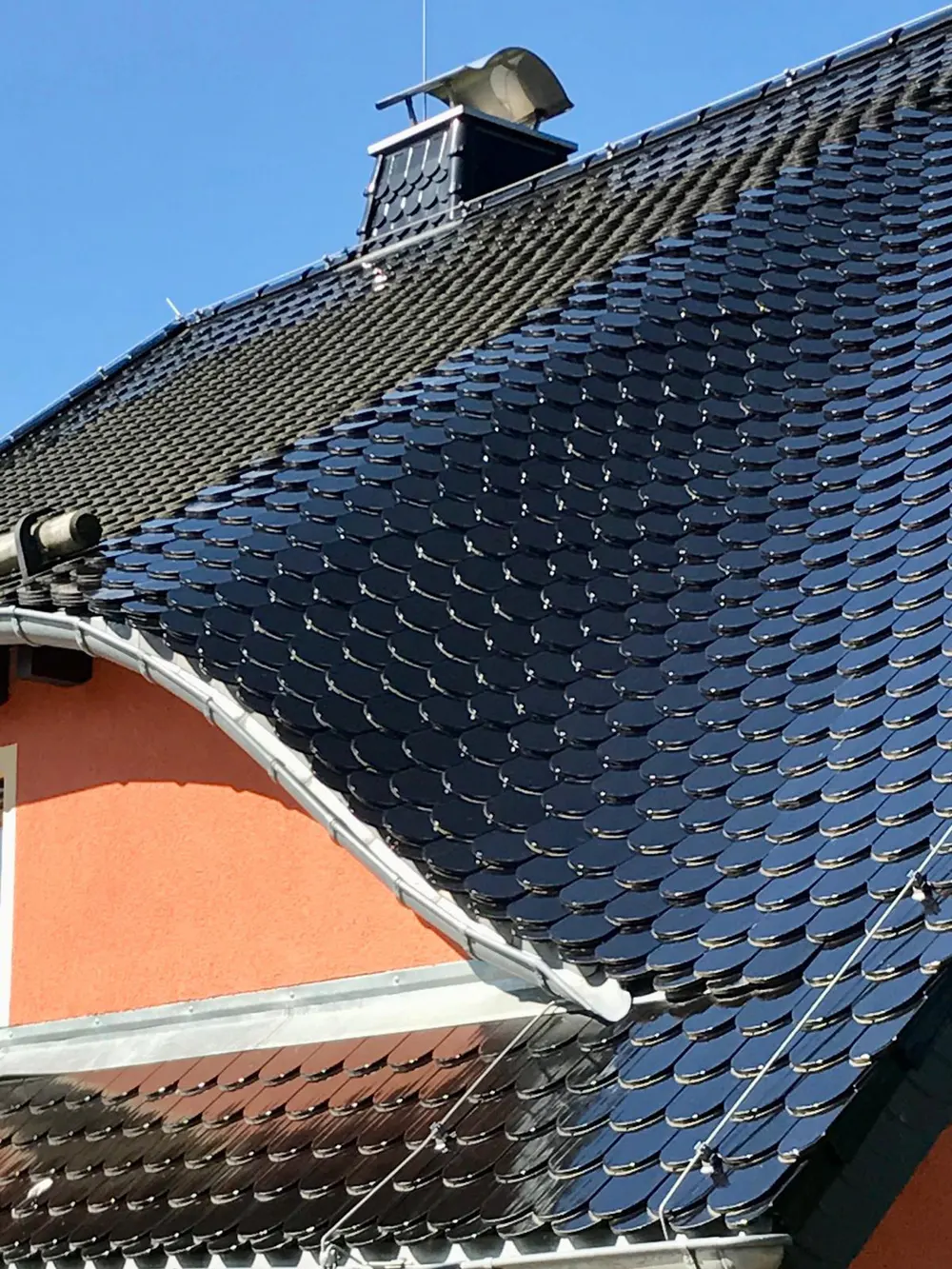 Dach eines Hauses mit glänzenden, schwarzen Schindeln, Regenrinne und Schornstein unter blauem Himmel.