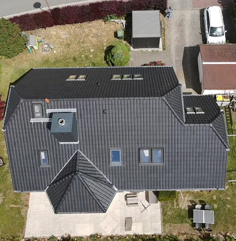 Dach eines Hauses mit schwarzen Ziegeln, Schornstein, Dachfenstern und Terrasse, von oben gesehen.