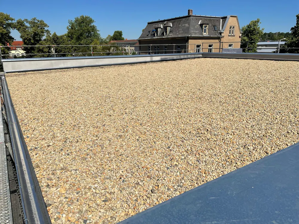 Kiesbedecktes Flachdach mit einem historischen Gebäude und Bäumen im Hintergrund unter blauem Himmel.