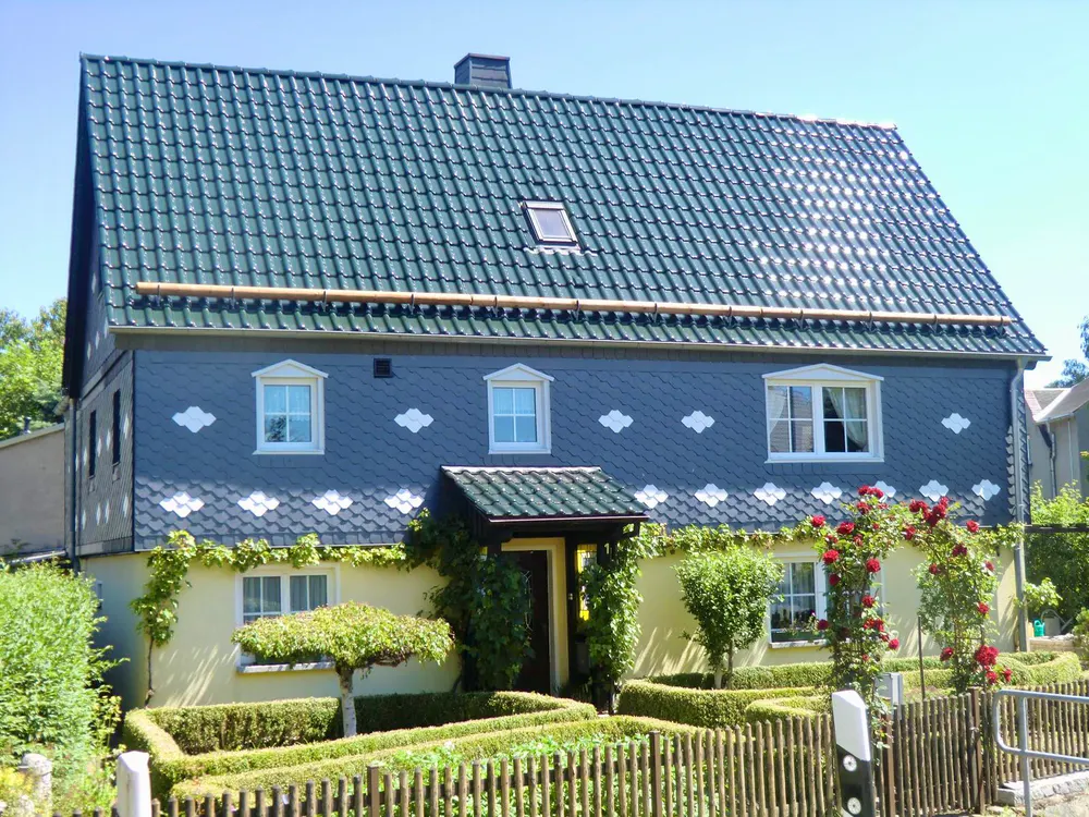 Blaues Schieferhaus mit gelber Fassade, grünem Dach, Giebel, Dachfenster, und einem Garten mit Rosen und Hecken.