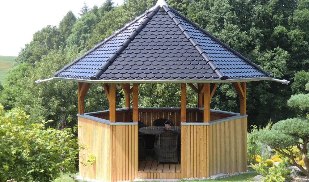 Holzpavillon mit sechseckigem, dunklem Ziegeldach, umgeben von grünen Bäumen und Sträuchern.