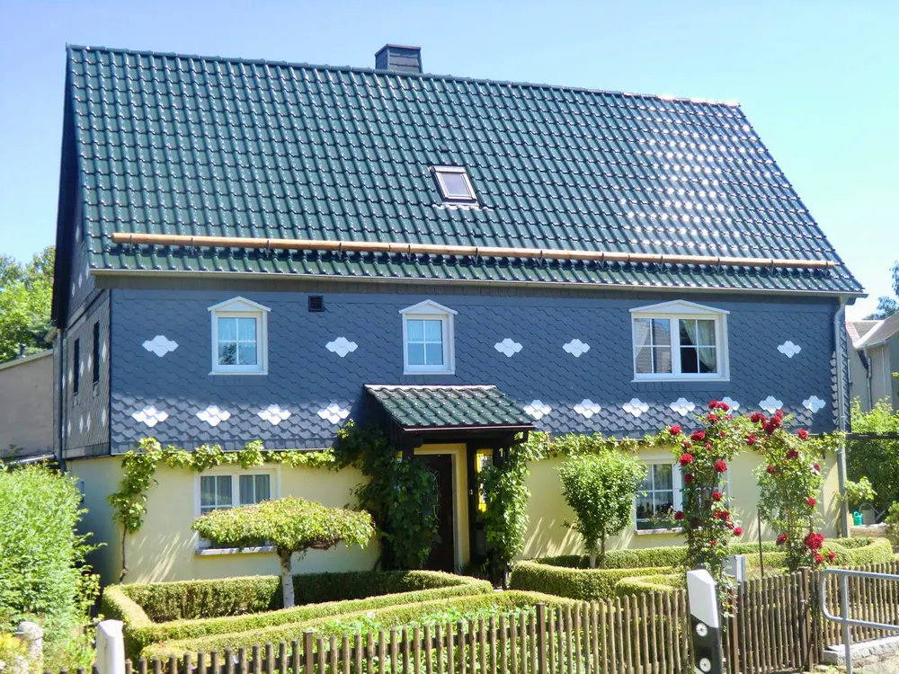 Ein zweistöckiges Haus mit dunkelblauem Schieferdach und Fassade, gelbem Erdgeschoss, umrankt von Kletterpflanzen und Rosen.