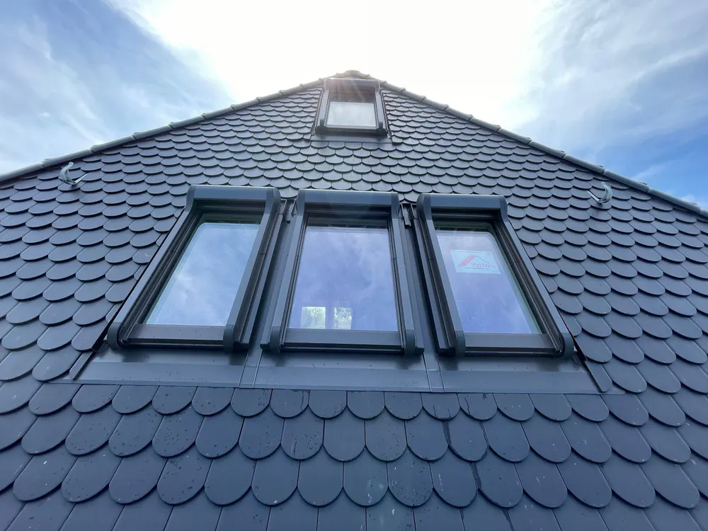 Drei Dachfenster mit schwarzen Rahmen und Rollläden, darüber ein weiteres kleines Dachfenster in einem grauen Schieferdach.