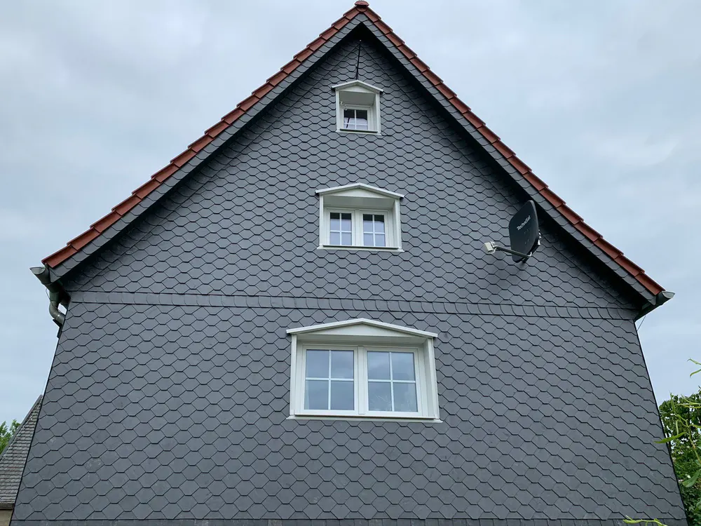 Giebel eines Hauses mit grauen Schieferplatten, drei weißen Fenstern und rotem Ziegeldach unter bewölktem Himmel.