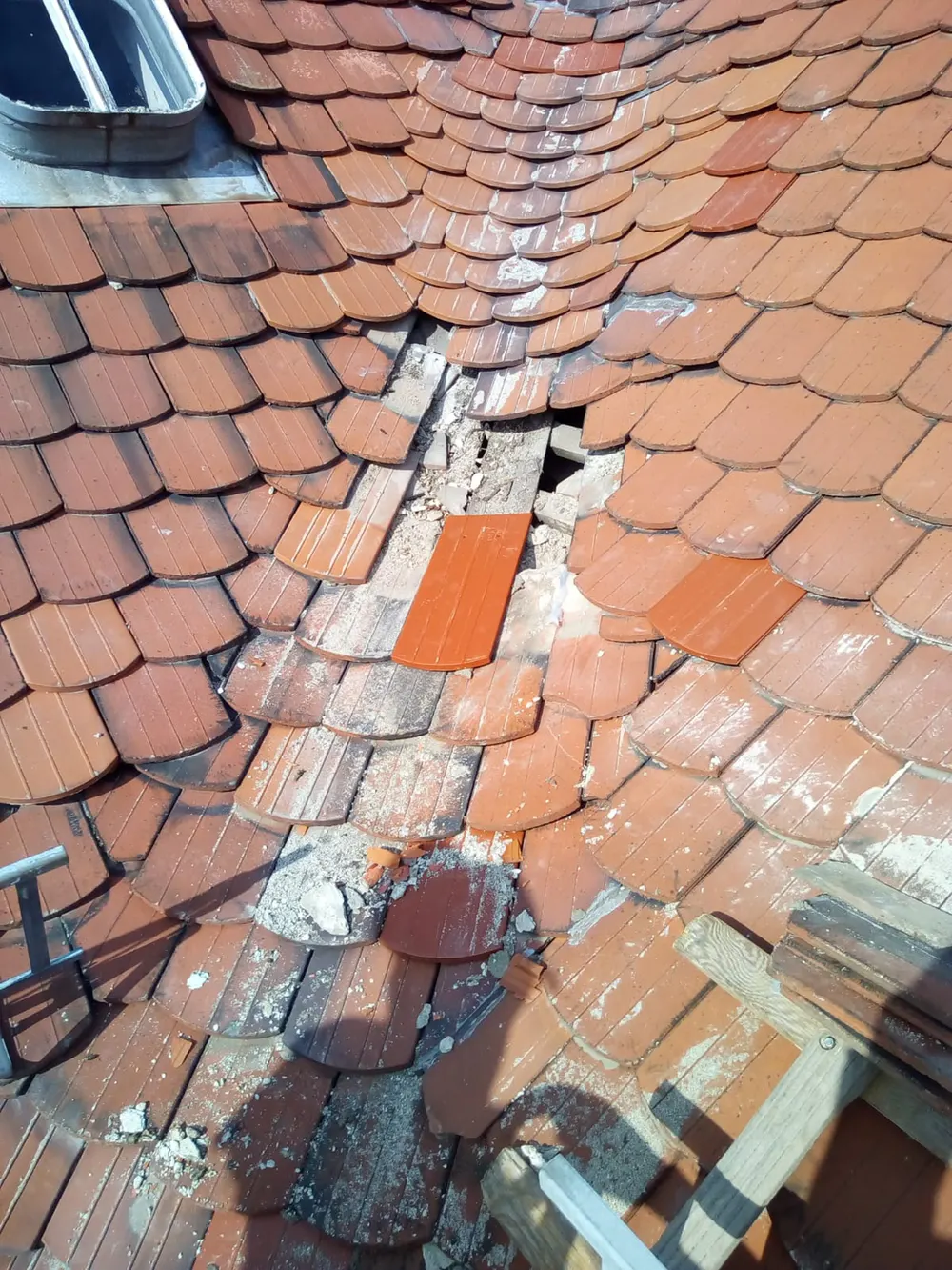 Beschädigtes Ziegeldach mit fehlenden und zerbrochenen Dachziegeln, darunter ein Dachfenster und eine Leiter.