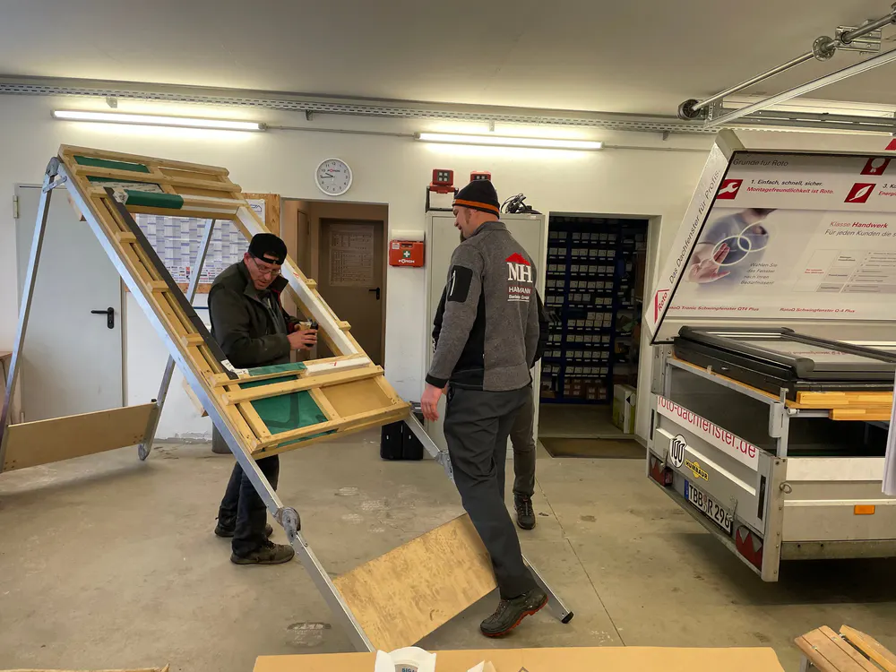 Zwei Männer montieren ein Dachfenster auf einem Holzrahmen in einer Werkstatt.