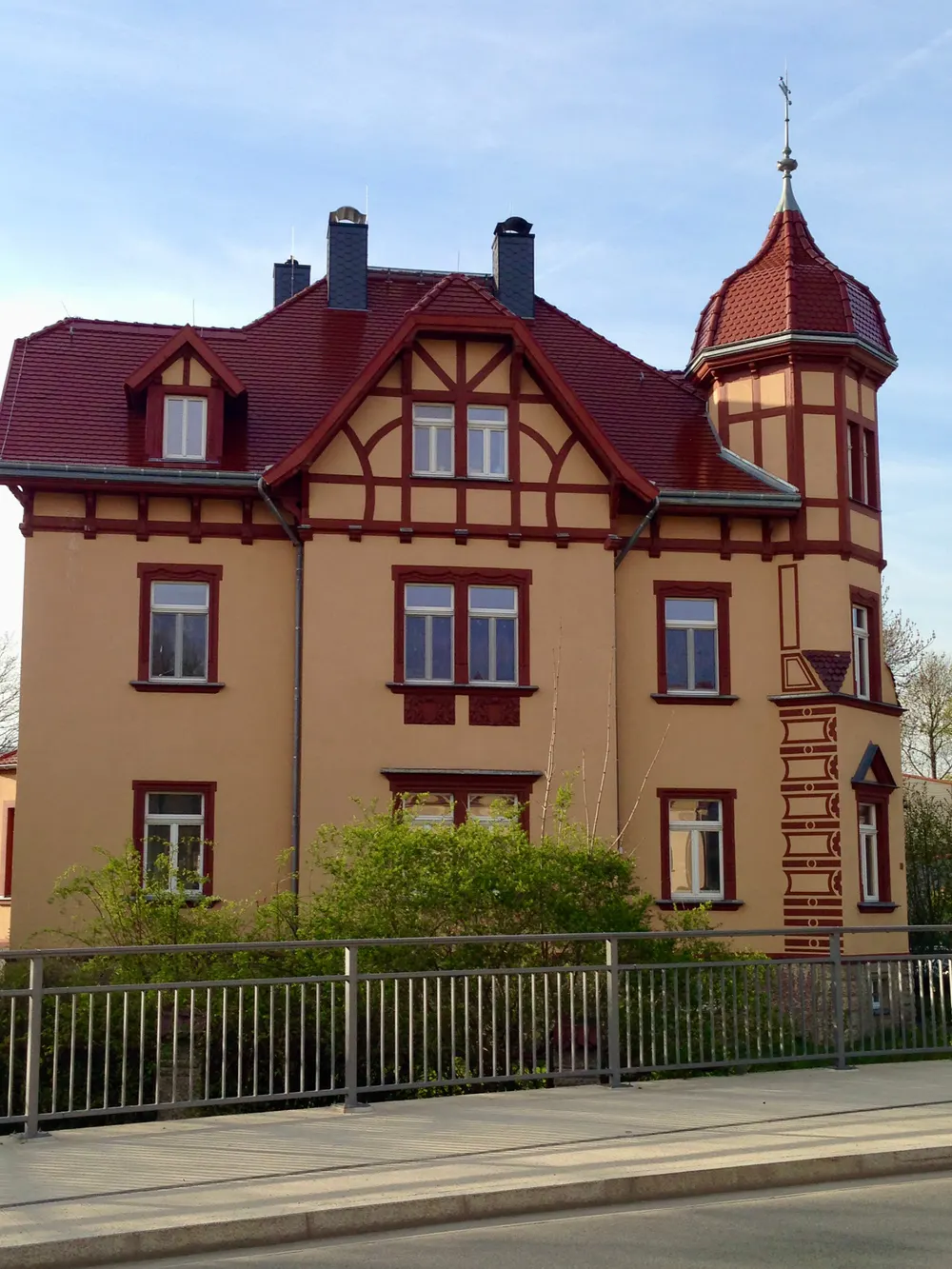Beiges Haus mit rotem Dach, Fachwerk-Giebel und einem Turm mit Wetterfahne, hinter einem Metallgeländer.