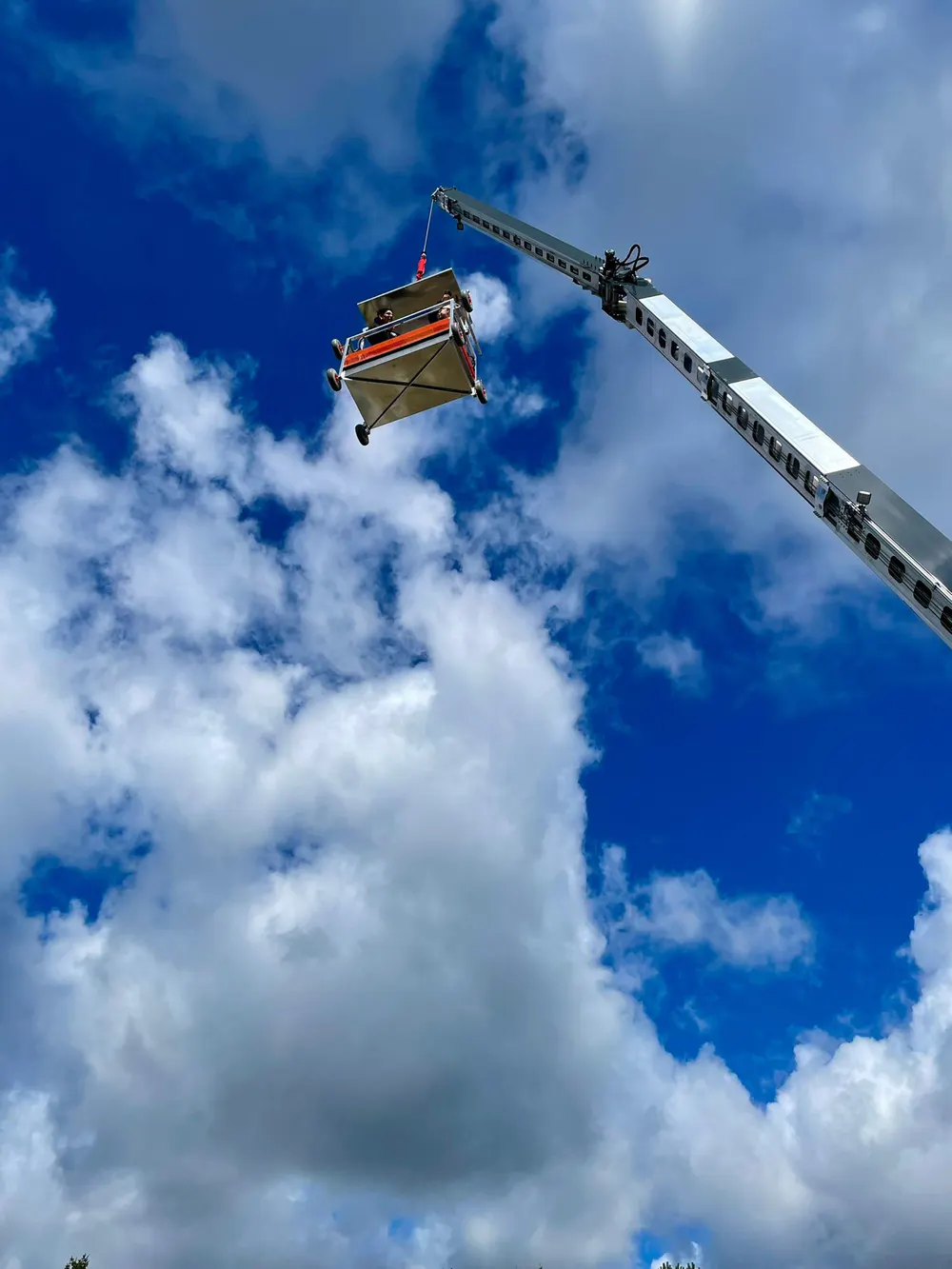 Ein Kranarm hebt eine orangefarbene Gondel mit Personen vor einem blauen Himmel mit weißen Wolken.