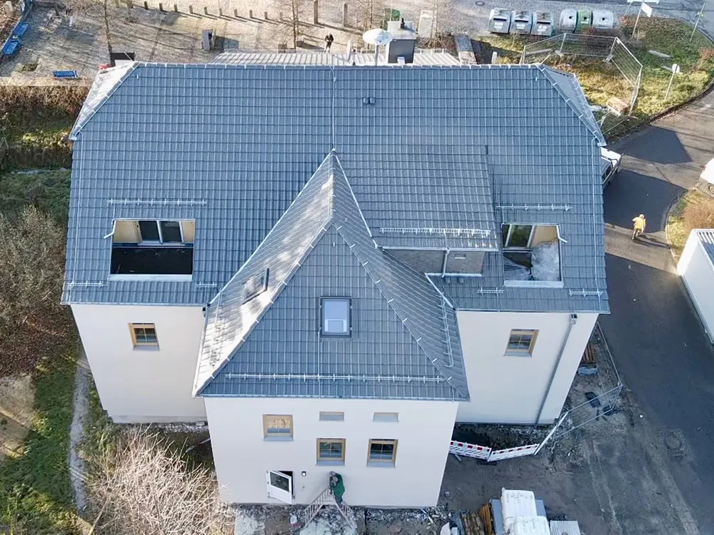 Drohnenaufnahme eines neuen Hauses mit grauem Ziegeldach, Gauben und einer Person vor der Haustür.