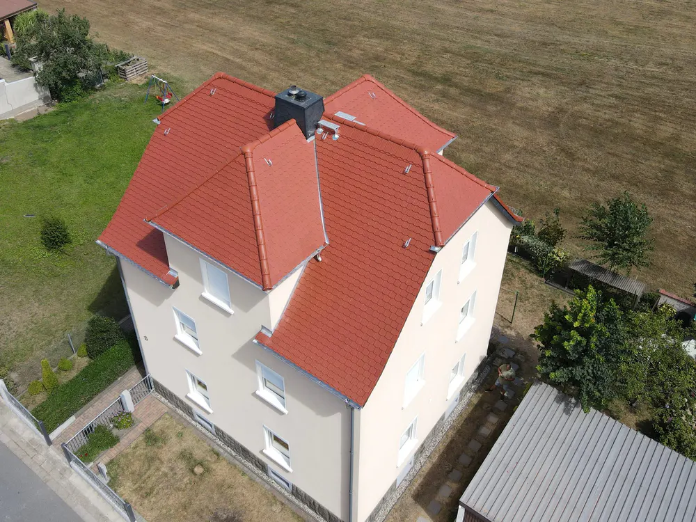 Luftaufnahme eines beigen Hauses mit neuem roten Dach und Schornstein, umgeben von grünen Wiesen und Bäumen.