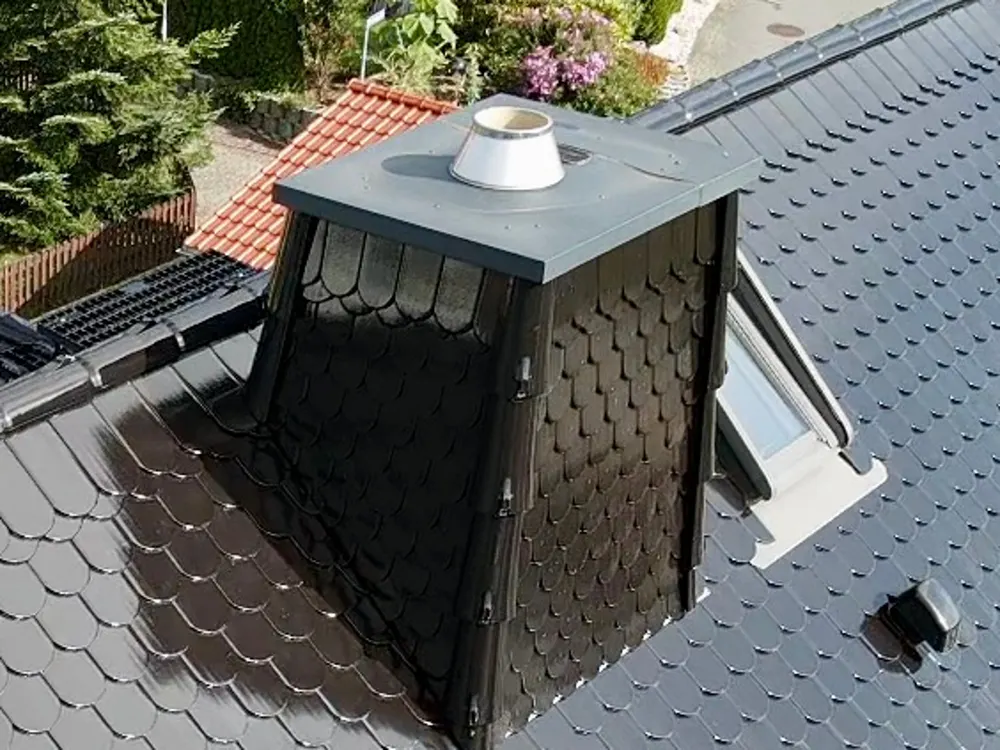 Dunkler Schornstein mit Fischschuppen-Verkleidung auf einem Schrägdach mit Dachfenster.