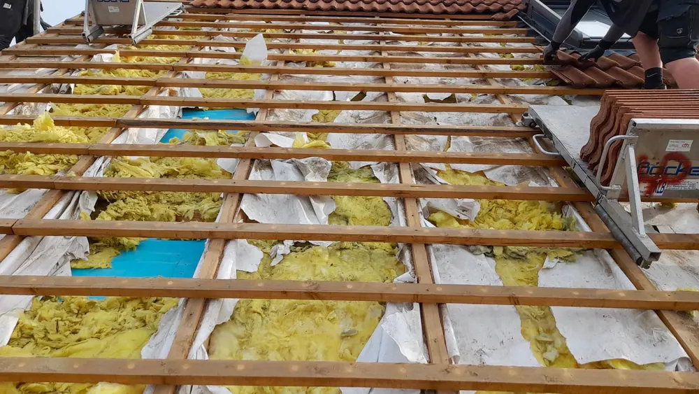 Dachstuhl mit Holzbalken, gelber Dämmung und blauer Folie, Dachdecker arbeitet oben rechts.