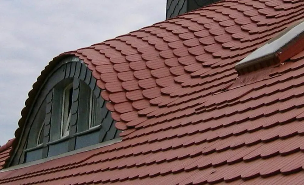 Rotes Schindeldach mit einem gewölbten Dachfenster, das von dunklen Schindeln umgeben ist.
