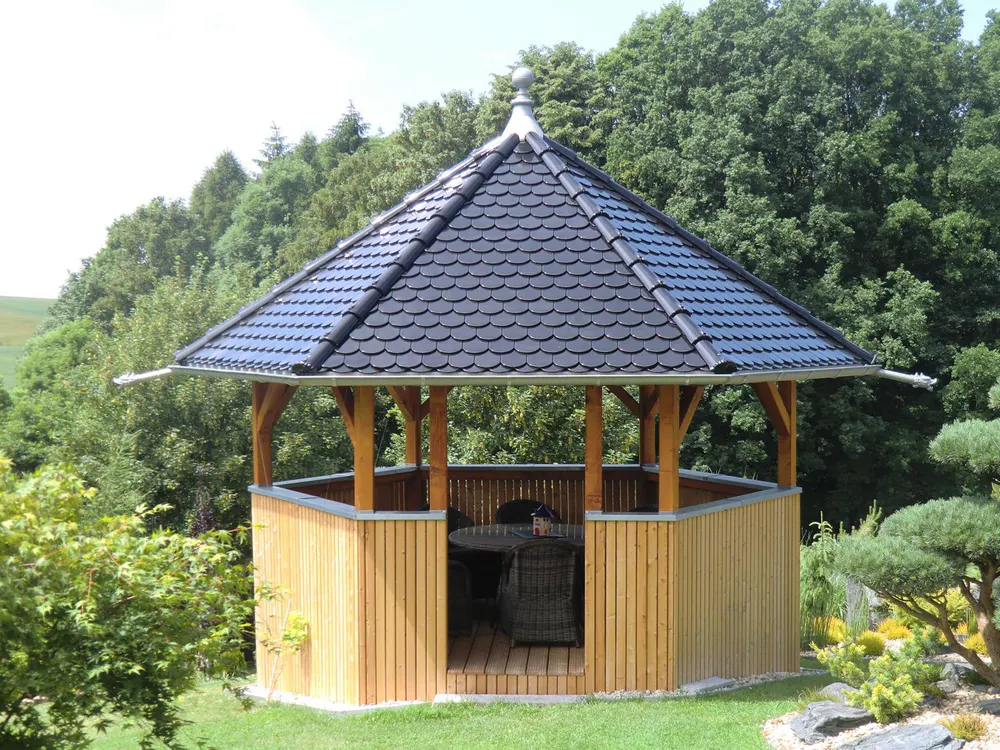 Holzpavillon mit schwarzem Ziegeldach und Holzboden in einem Garten mit Bäumen und Sträuchern.