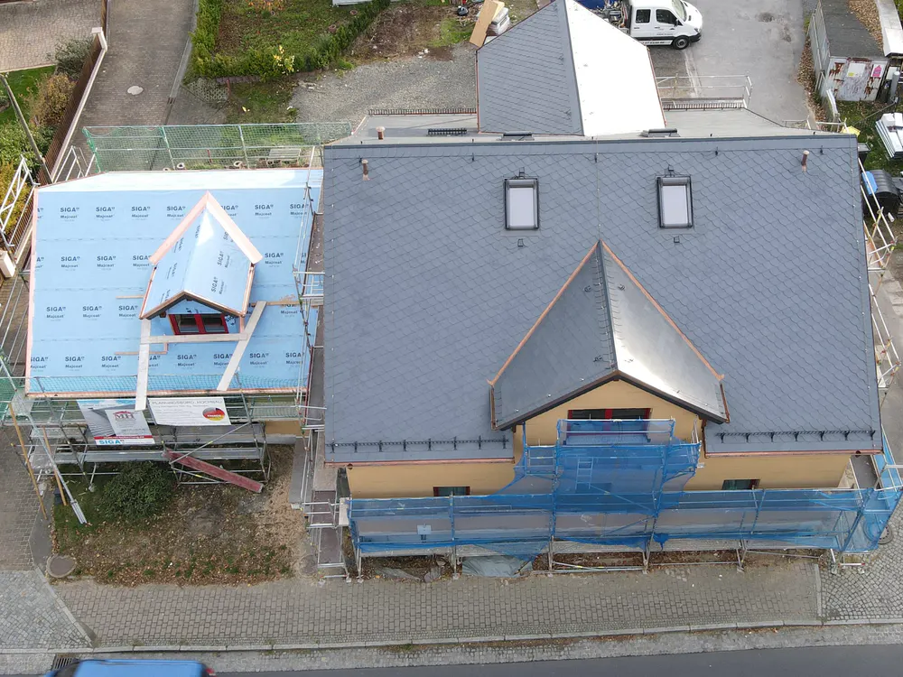 Draufsicht auf zwei Häuser im Bau; eines mit fertigem Schieferdach, das andere mit blauer Isolierung und Gerüst.