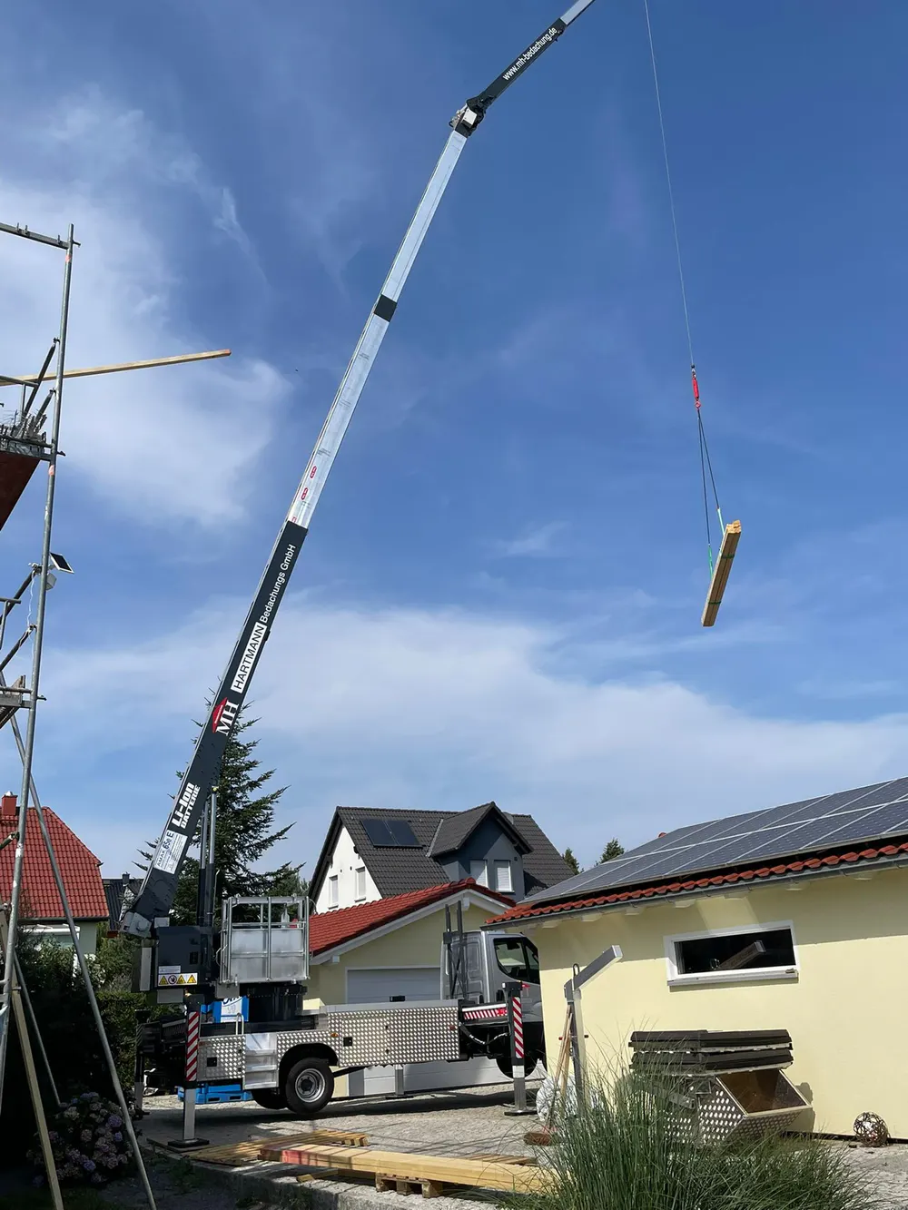 Ein Kran hebt ein Holzbrett über ein Haus mit Solarpaneelen unter blauem Himmel.