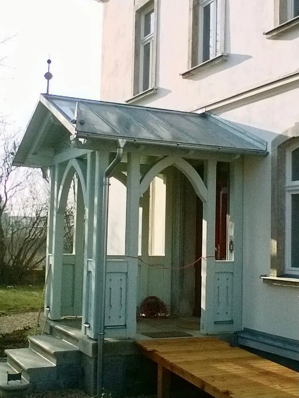Heller Holzvordach über einer Tür mit Stufen und einer Holzrampe, an einem weißen Haus.